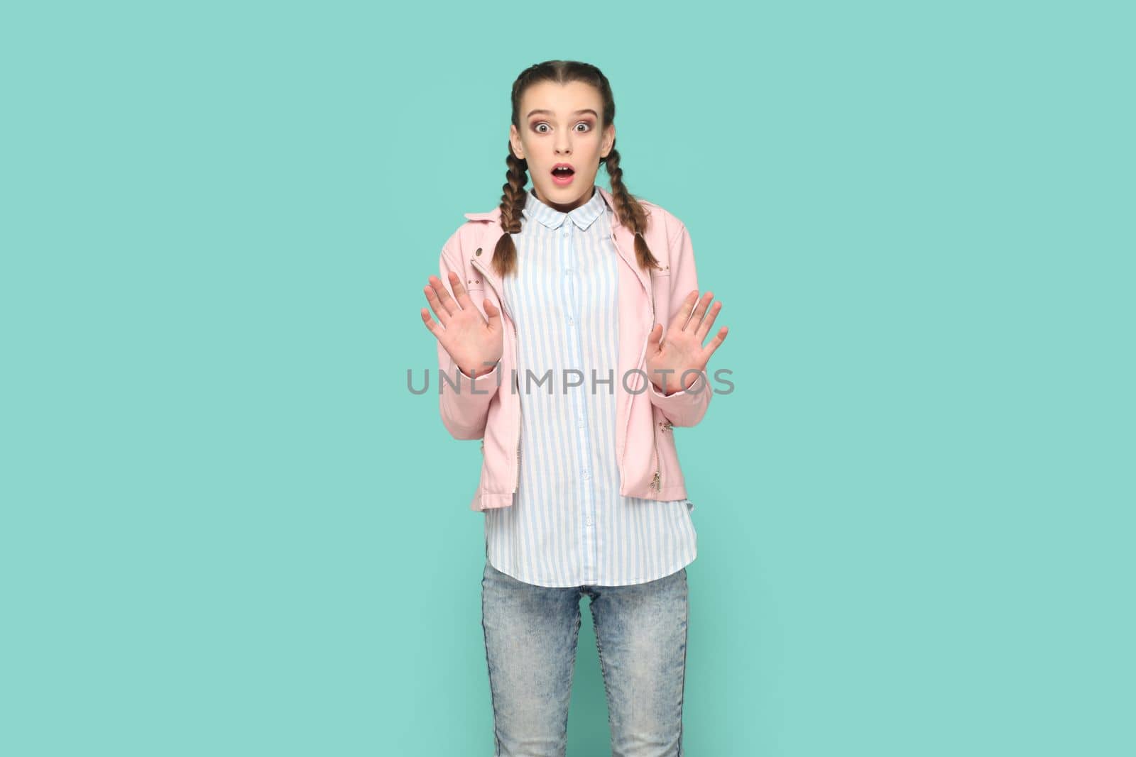 Portrait of scared young teenager girl with braids wearing pink jacket looking at camera with big eyes and open mouth, showing stop gesture. Indoor studio shot isolated on green background.
