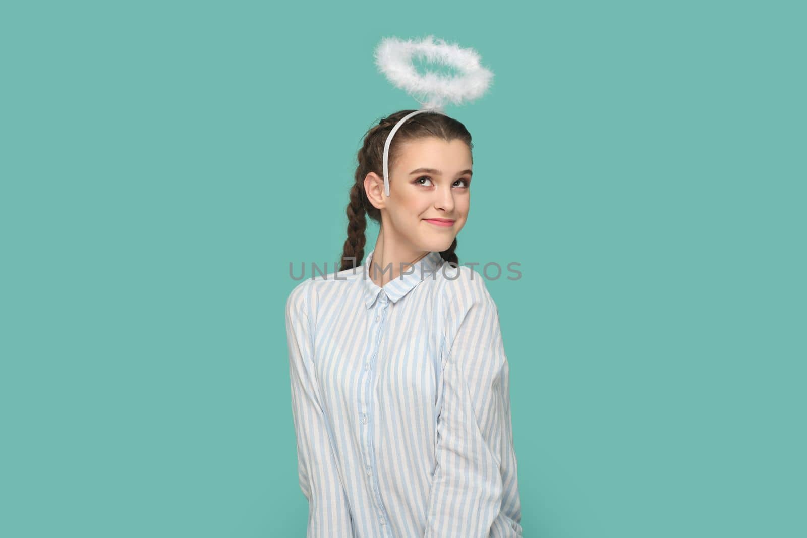 Portrait of pretty lovely friendly angelic teenager girl with braids wearing striped shirt and nimb over head, coquetry with boyfriend. Indoor studio shot isolated on green background.