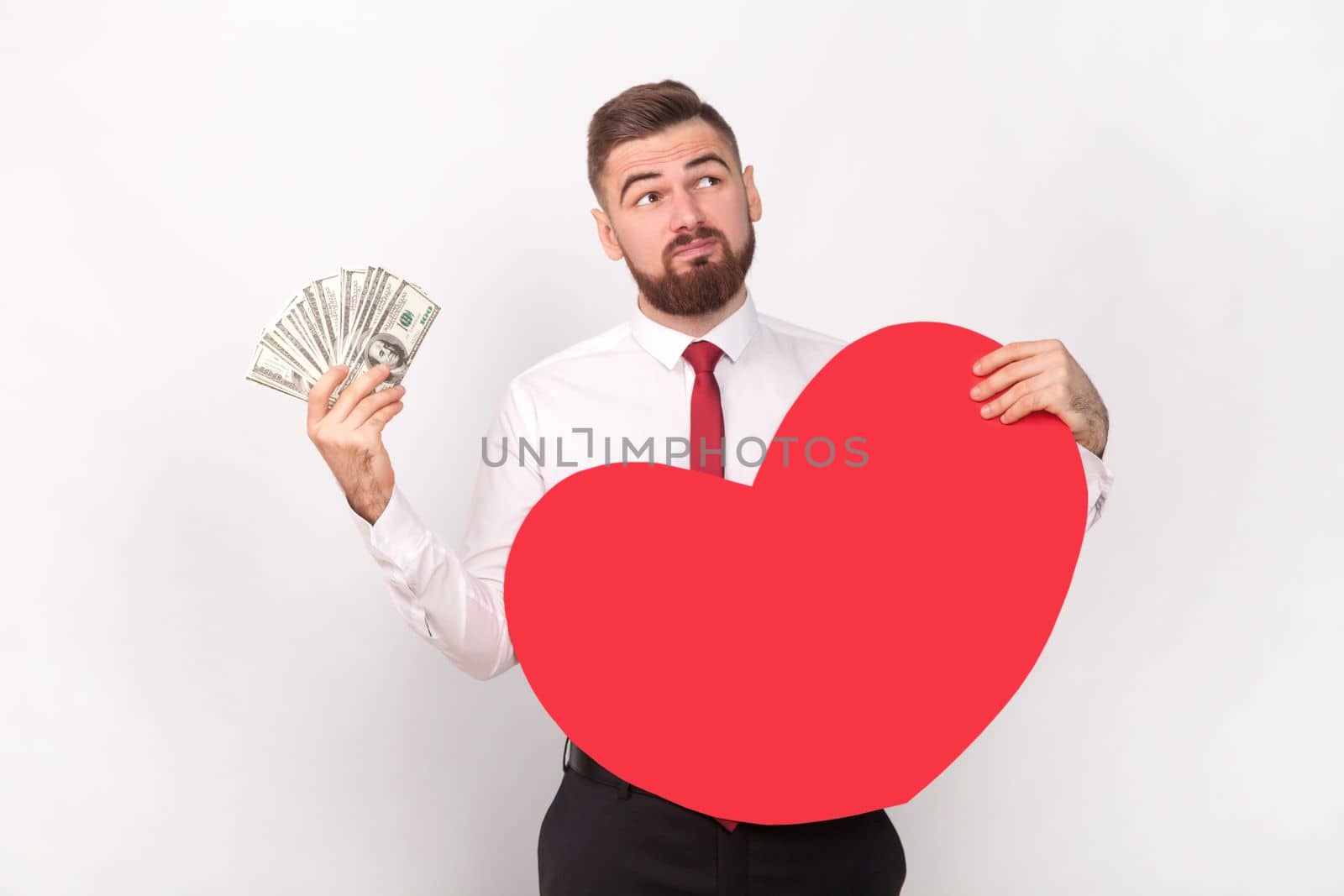 Portrait of puzzled confused pensive bearded man wearing white shirt and tie, holding big dollar banknotes and big red heart, looking away. Indoor shot isolated on gray background.