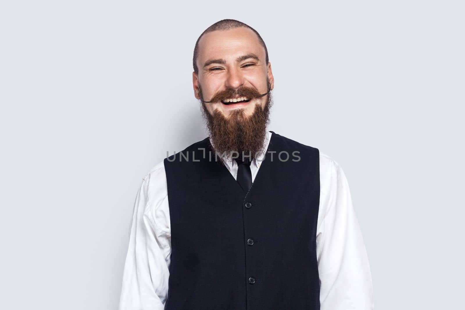 Portrait of excited attractive young adult man with beard and mustache standing and laughing, hearing funny jokes, looking at camera. Indoor studio shot isolated on gray background.