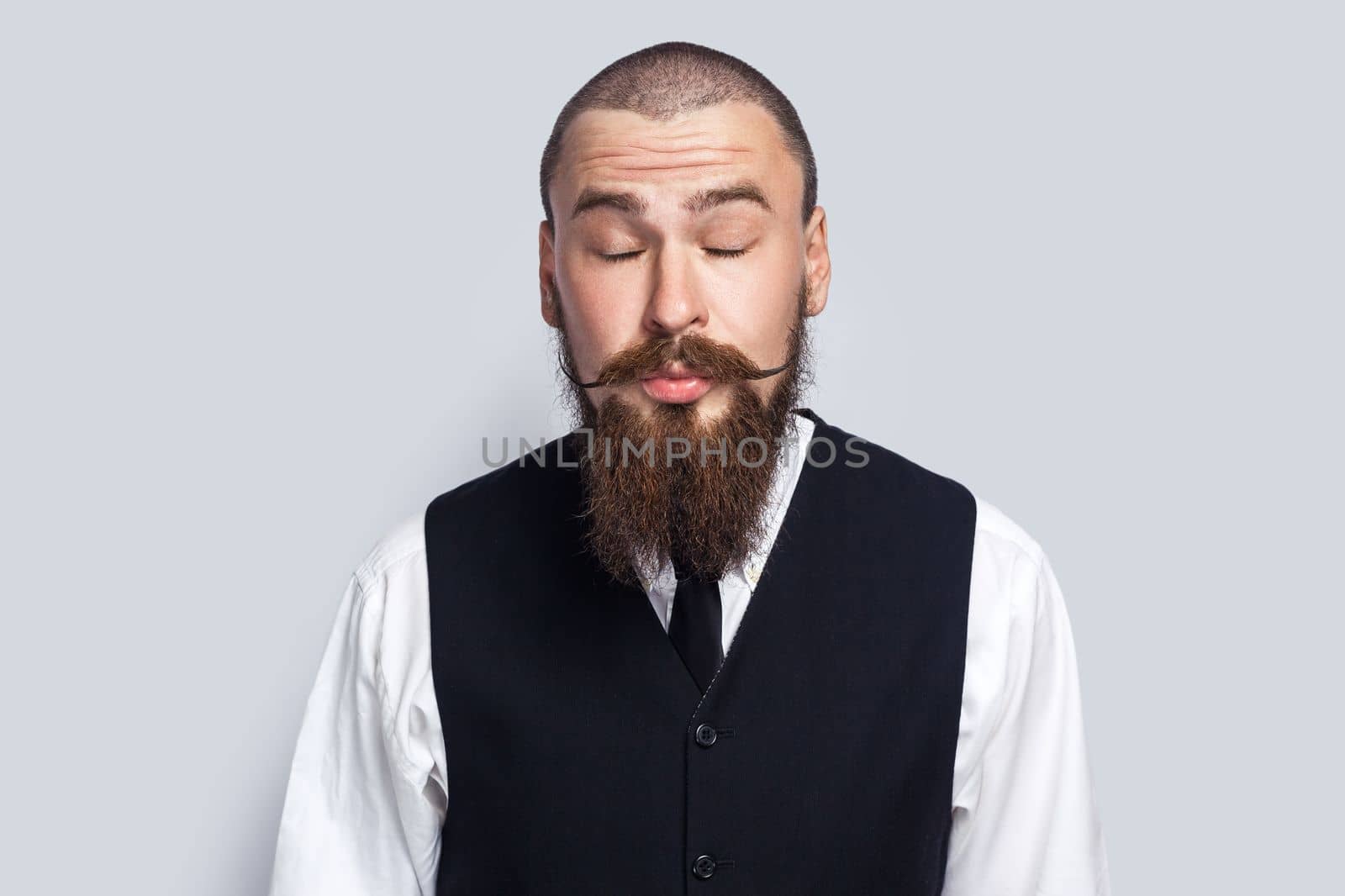 Portrait of shy flirting man with beard and mustache standing with closed eyes, sending air kisses, demonstrating his romantic feelings. Indoor studio shot isolated on gray background.