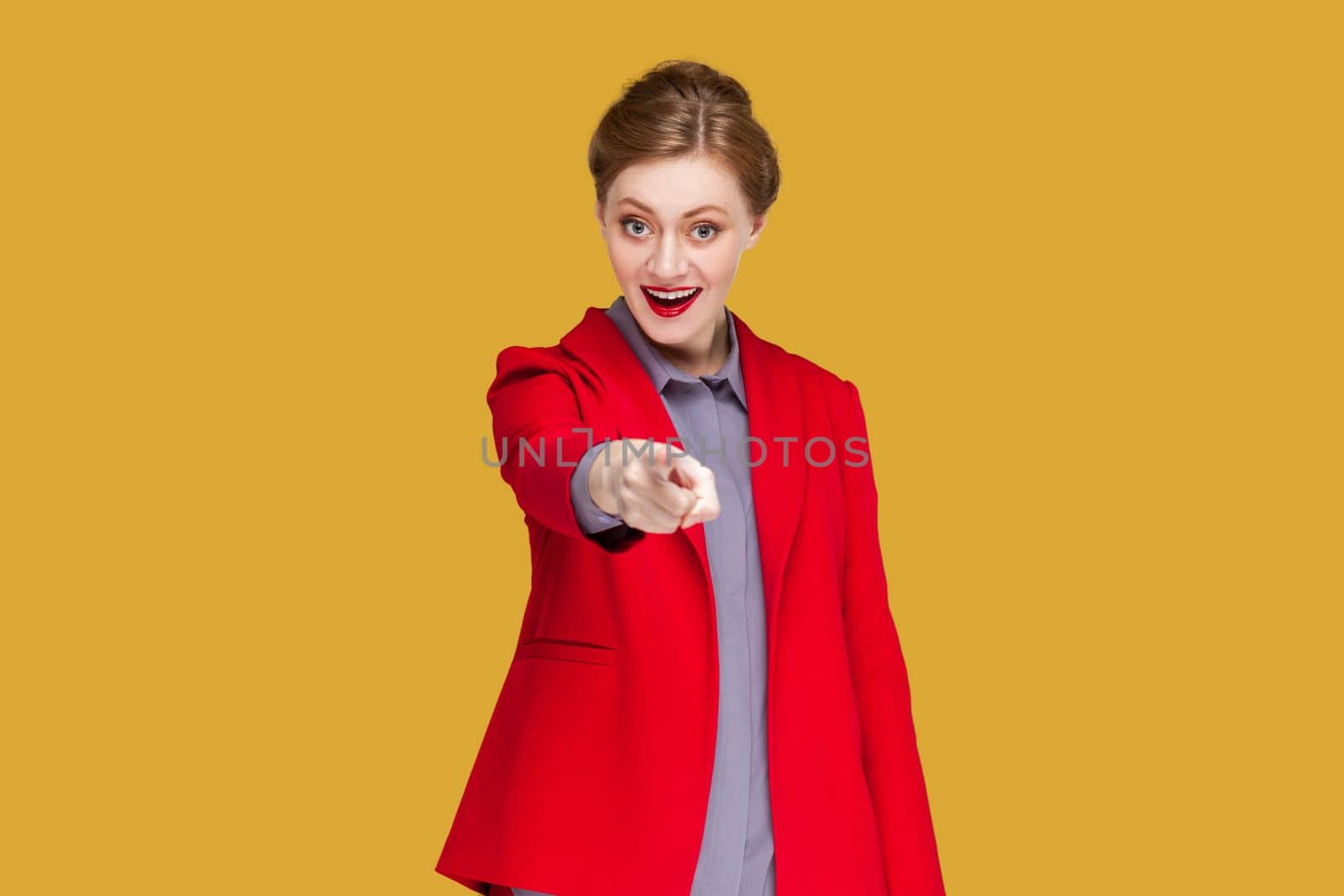 Portrait of happy excited woman with red lips standing pointing fore finger to camera, choosing you, expressing positive emotions, wearing red jacket. Indoor studio shot isolated on yellow background.