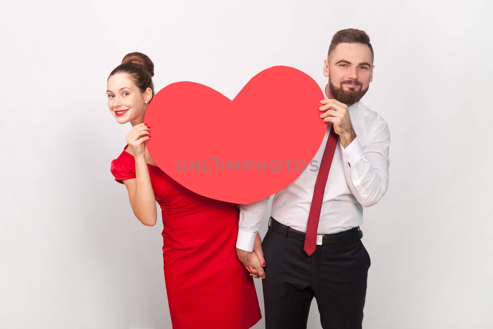 Portrait of smiling satisfied man in white shirt and woman in red dress standing together, peeping out from big heart, expressing love. Indoor studio shot isolated on gray background.