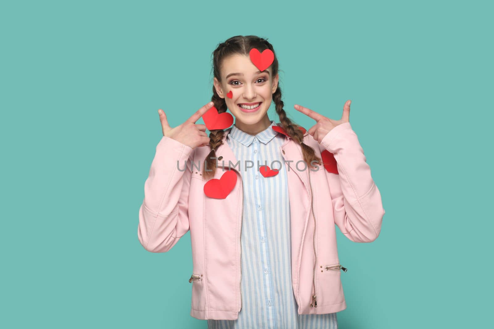 Portrait of romantic smiling teenager girl with braids wearing pink jacket standing pointing at little red heart on her body, expressing happiness. Indoor studio shot isolated on green background.