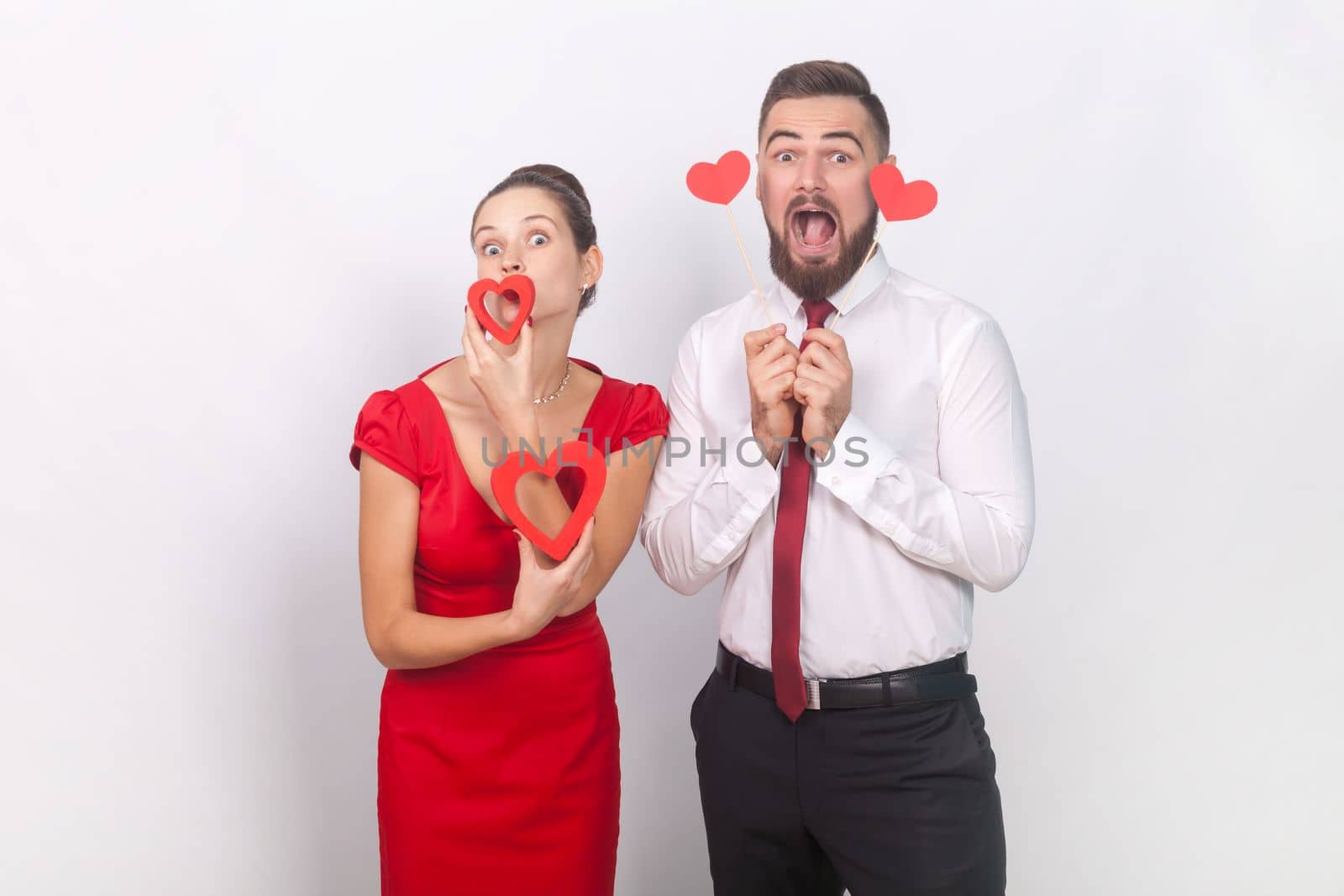 Portrait of funny screaming man in white shirt and woman in red dress with pout lips standing together, holding hear figures. Indoor studio shot isolated on gray background.