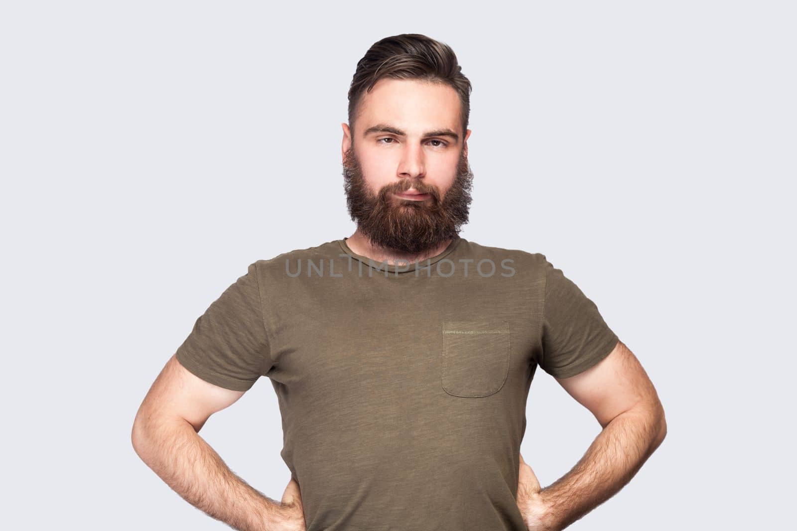 Portrait of bearded man wearing dark green T-shirt looking cross-eyed with stupid facial expression, fooling around, keeps hands on hips. Indoor studio shot isolated on light gray background.