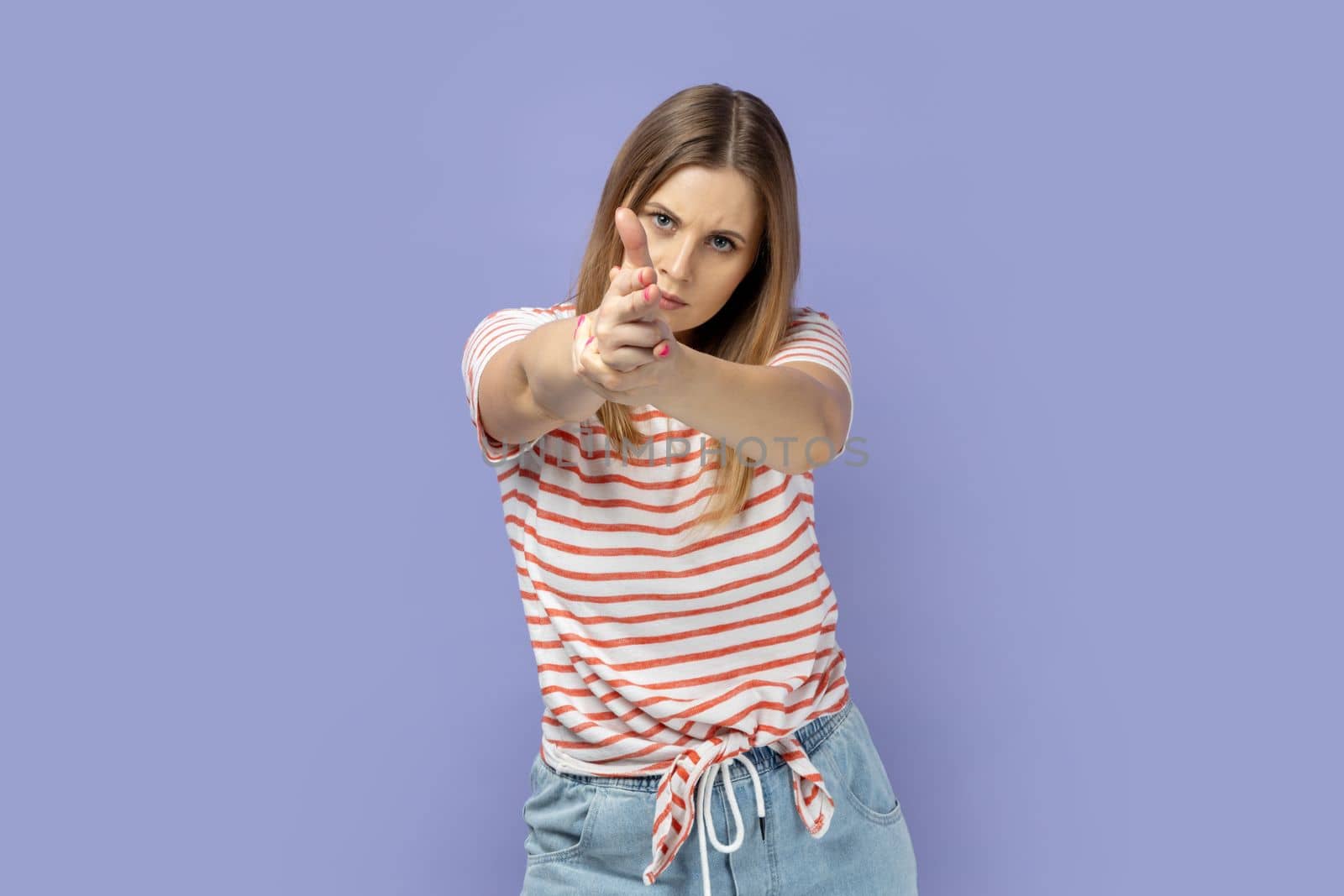 Portrait of serious brave blond woman wearing striped T-shirt holding fingers pretending holding gun, aiming enemy, self-defense, looking at camera. Indoor studio shot isolated on purple background.