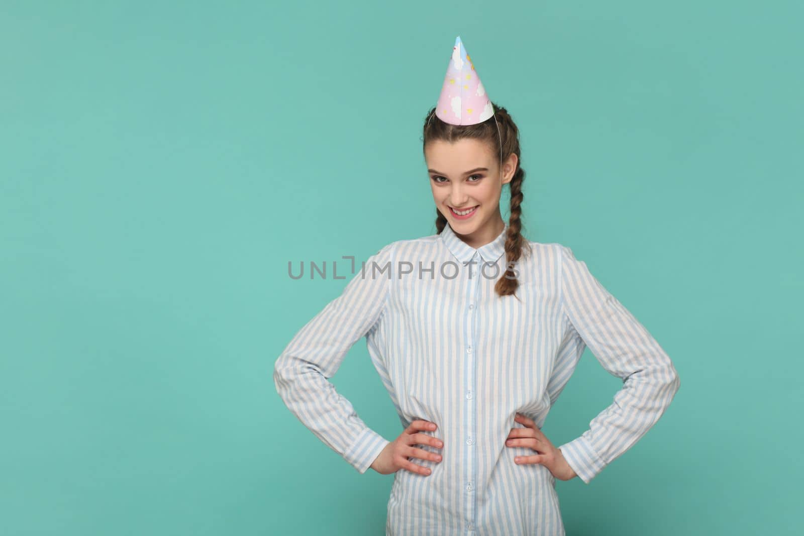 Portrait of happy festive teenager girl with braids wearing striped shirt and party cone, celebrating her birthday, keeps hands on hips. Indoor studio shot isolated on green background.