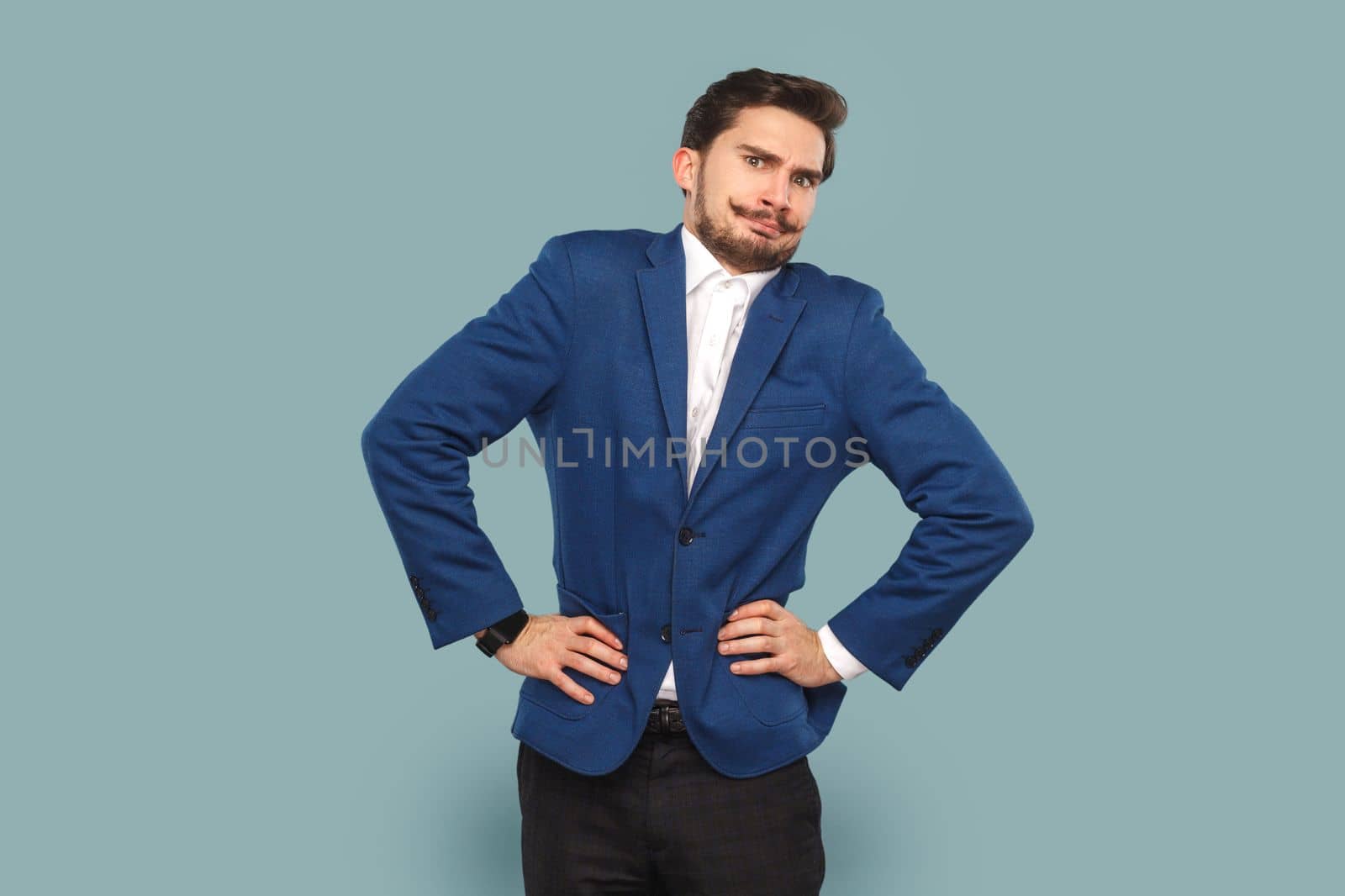 Portrait of funny angry man with mustache standing with hands on hips, looking at camera with frowning face, wearing official style suit. Indoor studio shot isolated on light blue background.