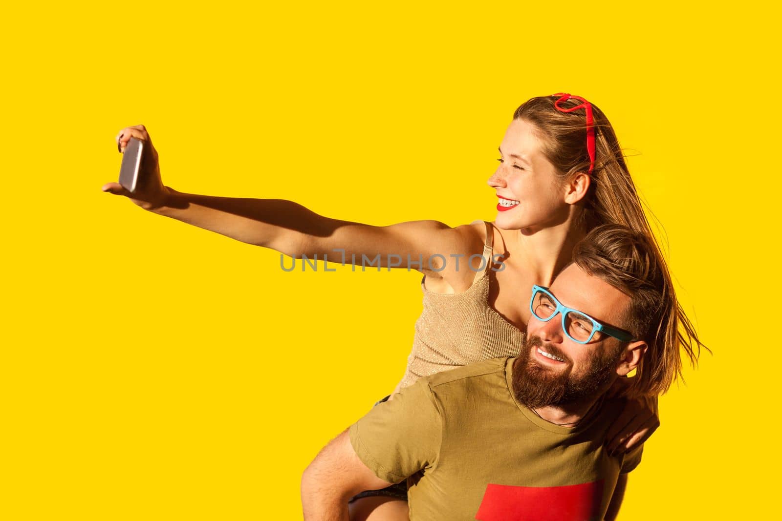 Portrait of happy funny positive man and woman making selfie on mobile phone, smiling happily, having fun together on date, piggyback. Indoor studio shot isolated on yellow background.