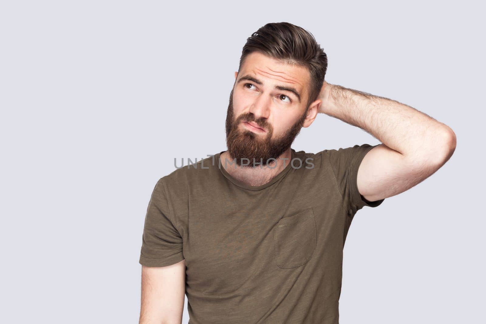 Portrait of pensive bearded man wearing dark green T-shirt touching head and thinking about important question, making hard decision. Indoor studio shot isolated on light gray background.