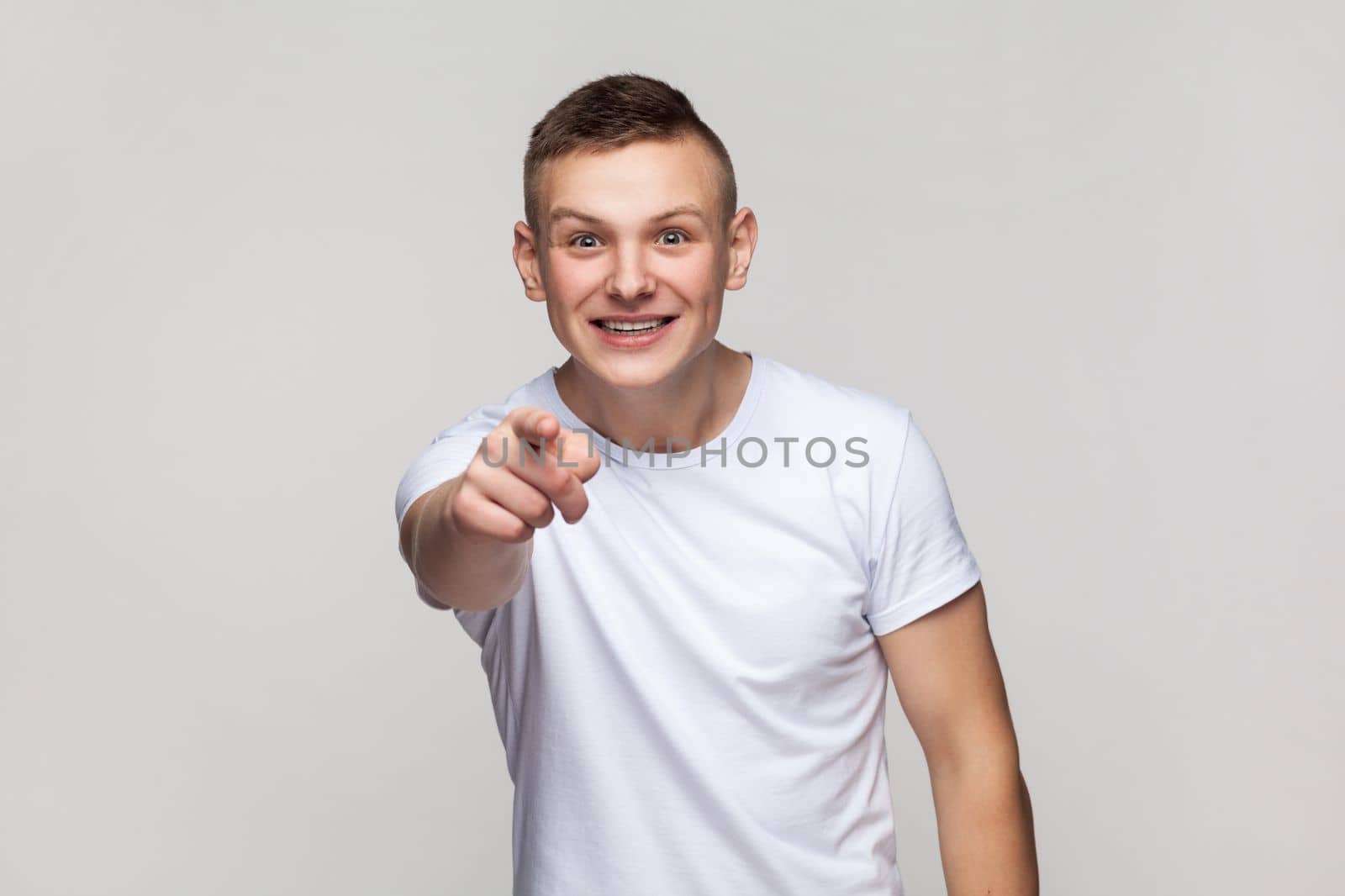 Portrait of astonished surprised young teenager boy wearing T-shirt looking at camera with big eyes, pointing at camera with finger, choosing you. Indoor studio shot isolated on gray background.