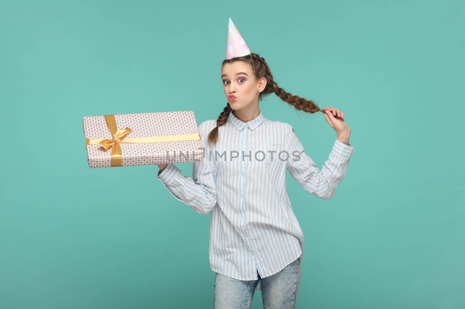 Flirting delighted attractive teenager girl with braids wearing striped shirt and party cone, holding her present on birthday, sending air kisses. Indoor studio shot isolated on green background.
