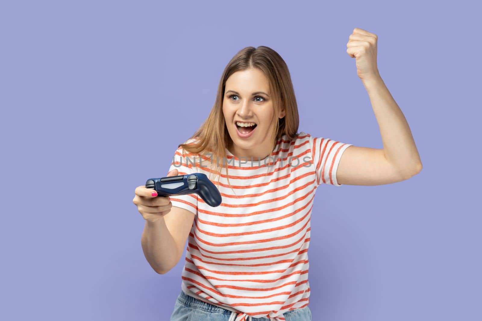 Portrait of excited amazed blond woman wearing striped T-shirt holding in hands gamepad joystick, clenched fist, showing yes gesture, winning. Indoor studio shot isolated on purple background.