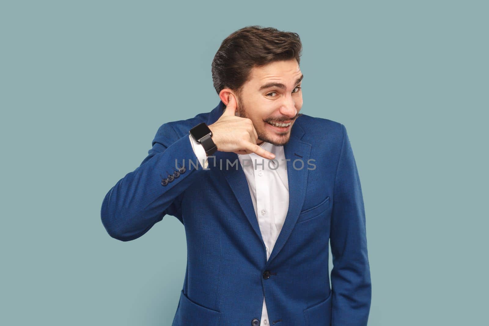 Portrait of happy positive man with mustache standing showing call me back gesture, flirting with beautiful woman, wearing official style suit. Indoor studio shot isolated on light blue background.