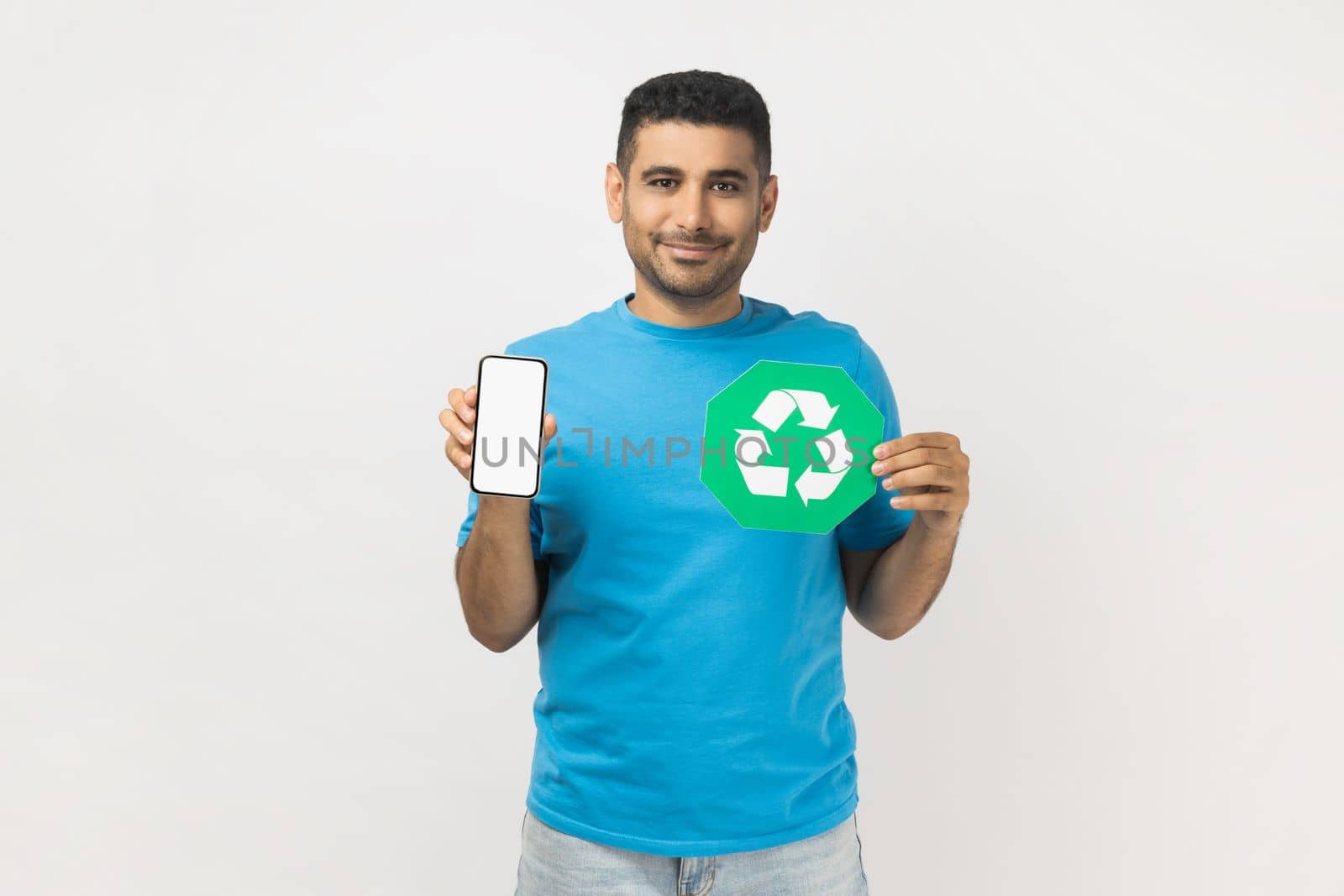 Portrait of happy unshaven man wearing blue T- shirt standing standing holding recycling sign and smart phone with blank screen for advertisement. Indoor studio shot isolated on gray background.