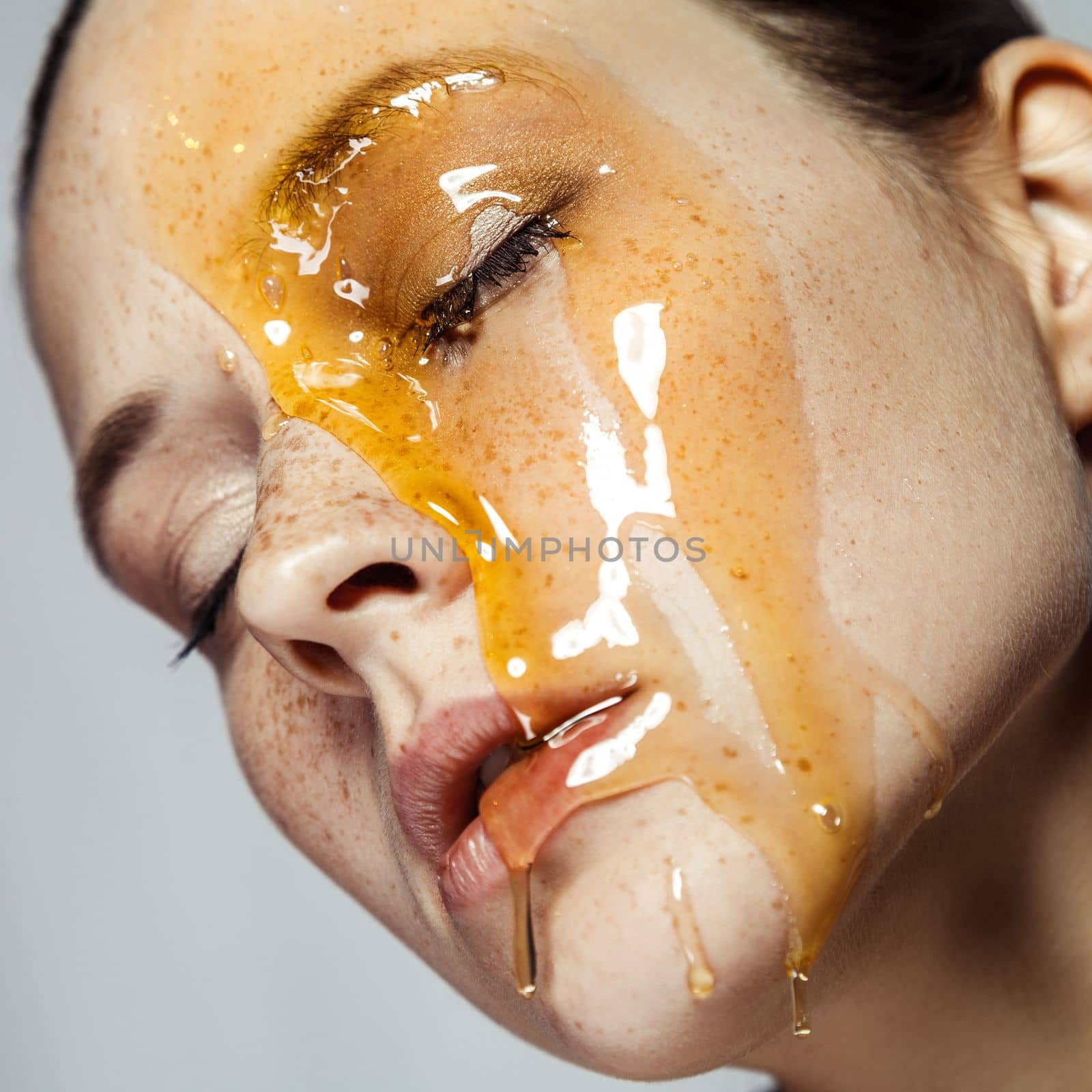 Closeup profile portrait of relaxed calm young brunette woman with freckles, standing with closed eyes, doing skin care procedures with honey mask. Indoor studio shot isolated on gray background.