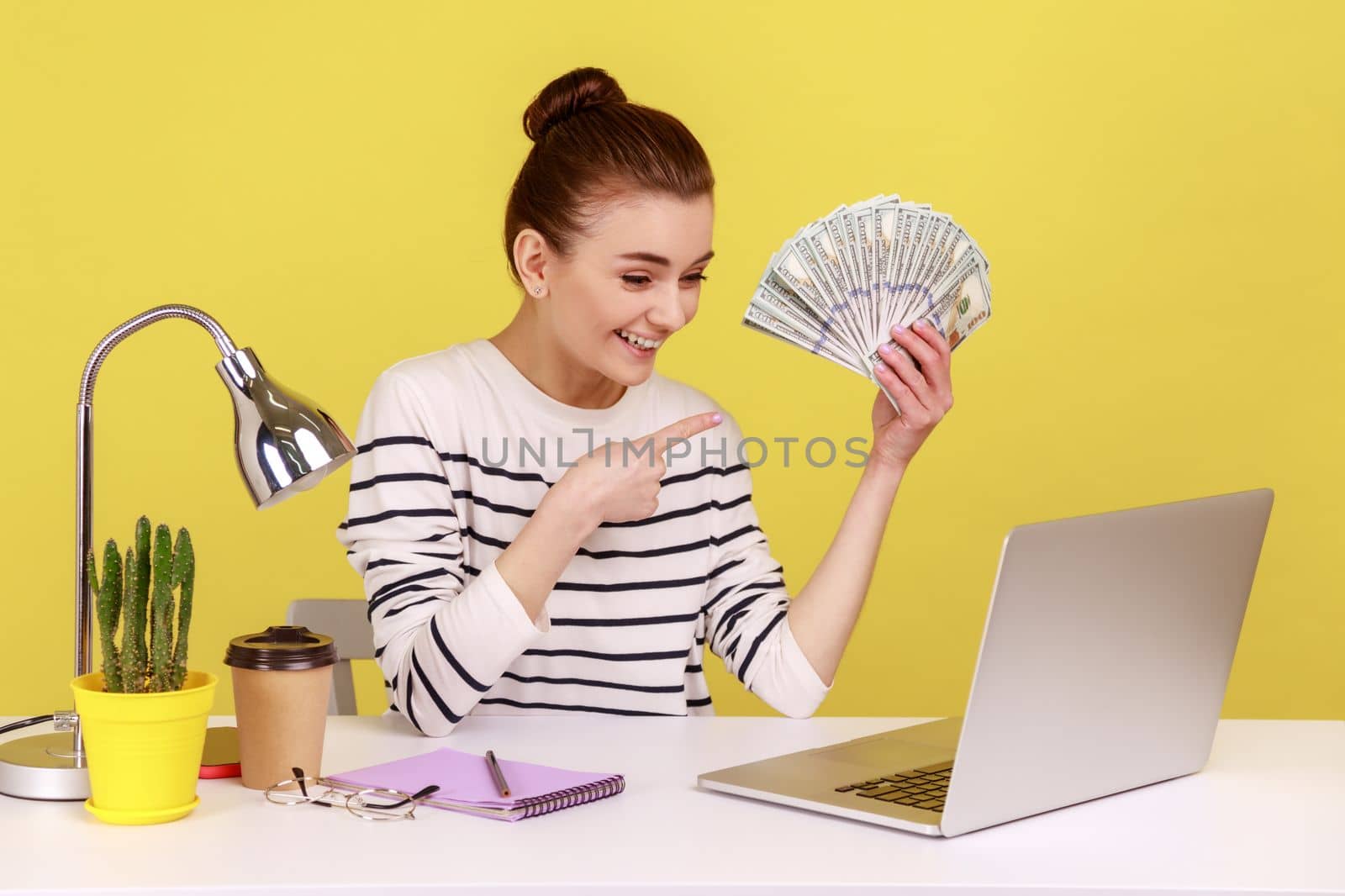 Positive young woman pointing finger at hundred dollar bills, holding sitting at workplace, looking at laptop, high salary, bonuses and perks. Indoor studio studio shot isolated on yellow background.