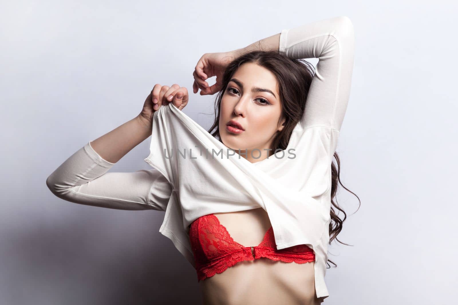 Portrait of cute beautiful young adult brunette woman with wavy hair taking off her shirt, showing her red underwear, expressing passion. Indoor shot isolated on gray background.