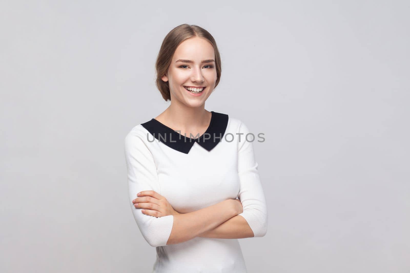 Portrait of smiling friendly woman wearing white dress standing with crossed arms, looking at camera with toothy smile, expressing positive emotions. Indoor studio shot isolated on gray background.
