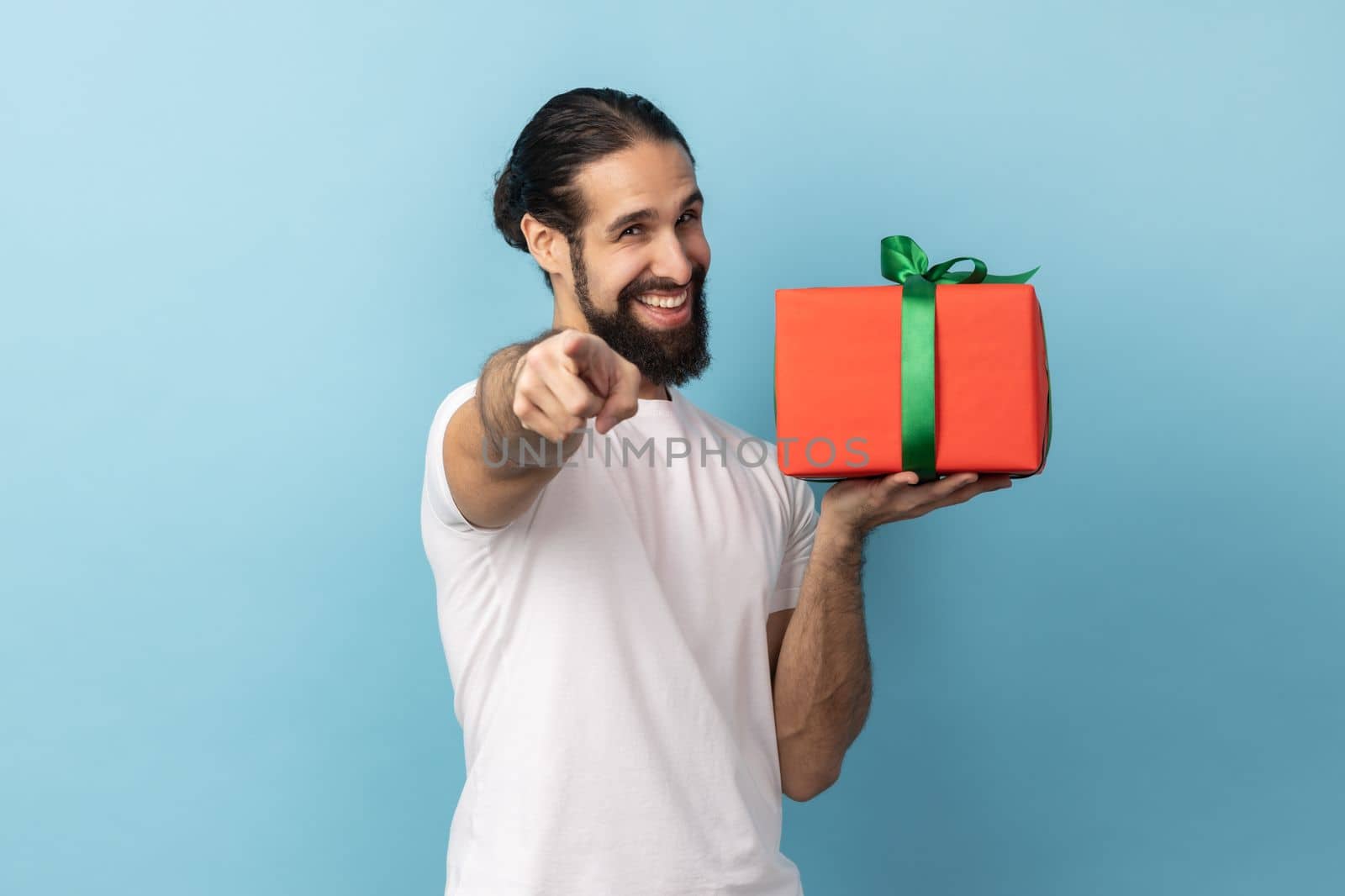 Portrait of positive man with beard wearing white T-shirt giving present and pointing finger to camera, sharing holiday gift, congratulating. Indoor studio shot isolated on blue background.