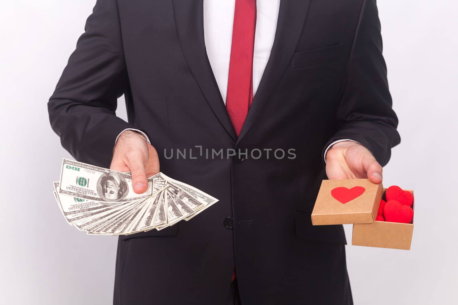 Portrait of unknown anonymous man wearing black official style suit, white shirt and red tie, holding little box with hearts and fan of dollar banknotes. Indoor studio shot isolated on gray background