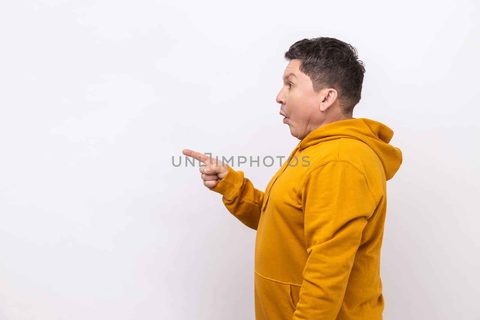 Side view portrait of shocked man pointing finger away at empty space for advertisement, looking aside with open mouth, wearing urban style hoodie. Indoor studio shot isolated on white background.