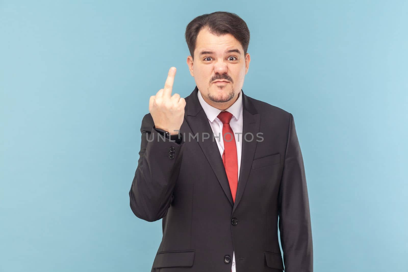 Portrait of serious aggressive man with mustache standing showing middle finger, expressing disrespect, wearing black suit with red tie. Indoor studio shot isolated on light blue background.