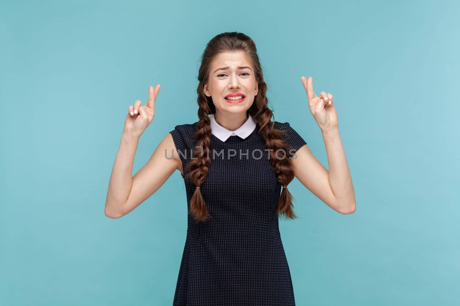 Portrait of attractive young woman with braids makes wish, frowning face, crosses fingers hopes for better, wearing black dress. woman Indoor studio shot isolated on blue background.