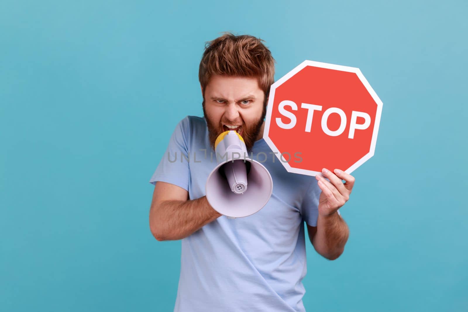 Portrait of aggressive angry bearded man looking at camera, screaming in loud speaker, holding red stop sign and megaphone in his hands. Indoor studio shot isolated on blue background.