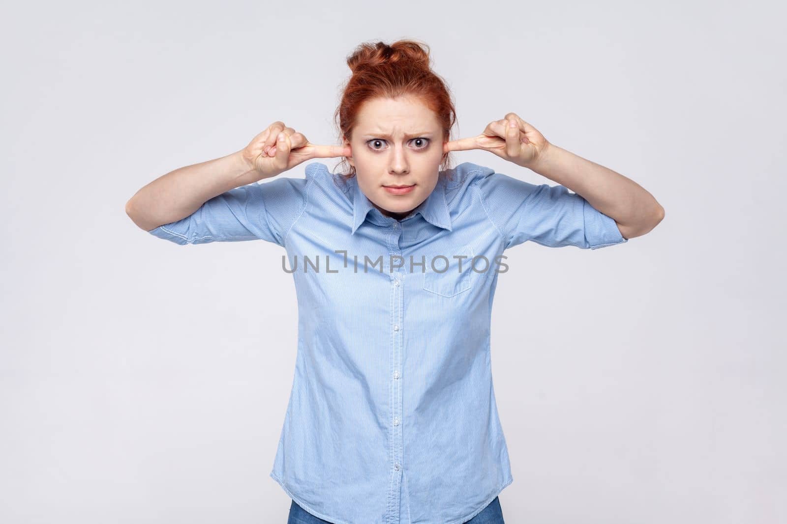 Don't want to listen. Portrait of annoyed redhead woman wearing blue shirt irritated by loud noise covering ears and grimacing in pain. Indoor studio shot isolated on gray background.