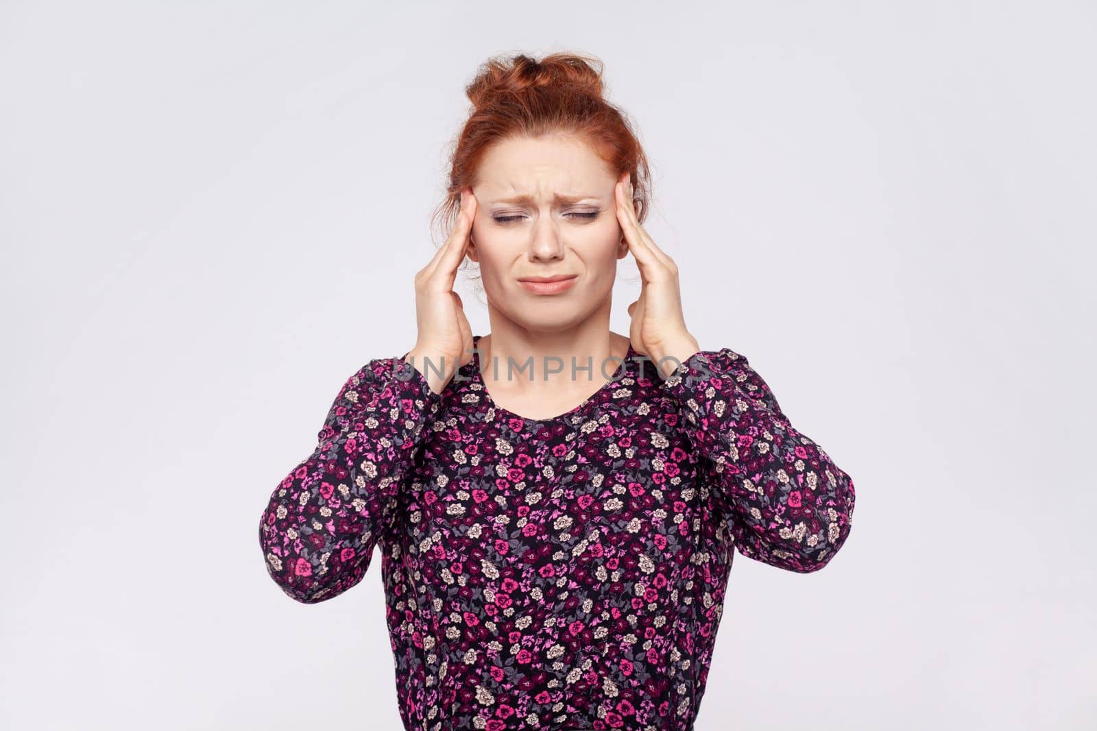 Portrait of unhealthy sick young adult redhead woman wearing dress massaging temples, suffering from headache, migraine, feels unwell. Indoor studio shot isolated on gray background.