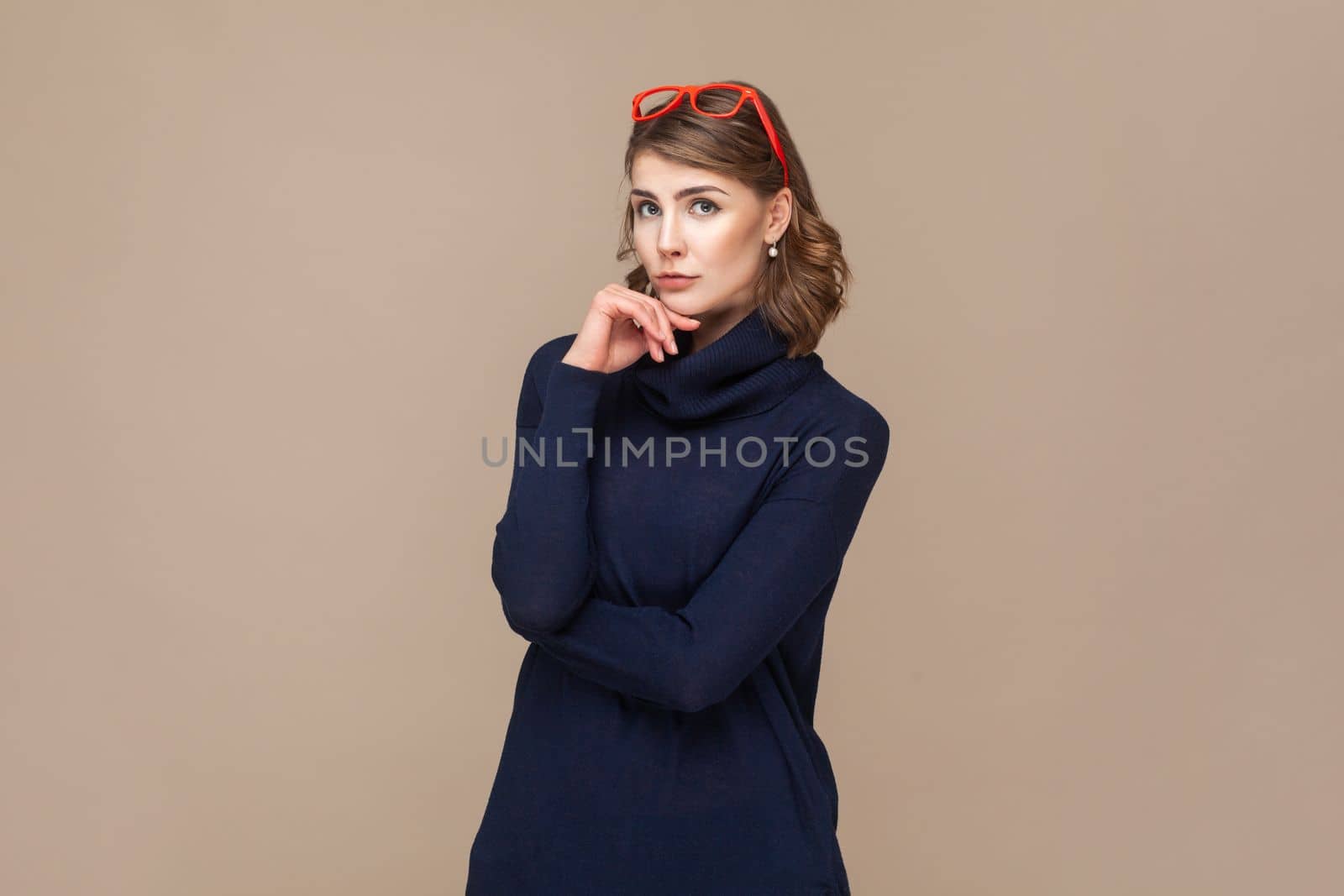 Portrait of pensive thoughtful woman with wavy hair in red red glasses on her head holding her chin, thinking about future. Indoor studio shot isolated on light brown background.