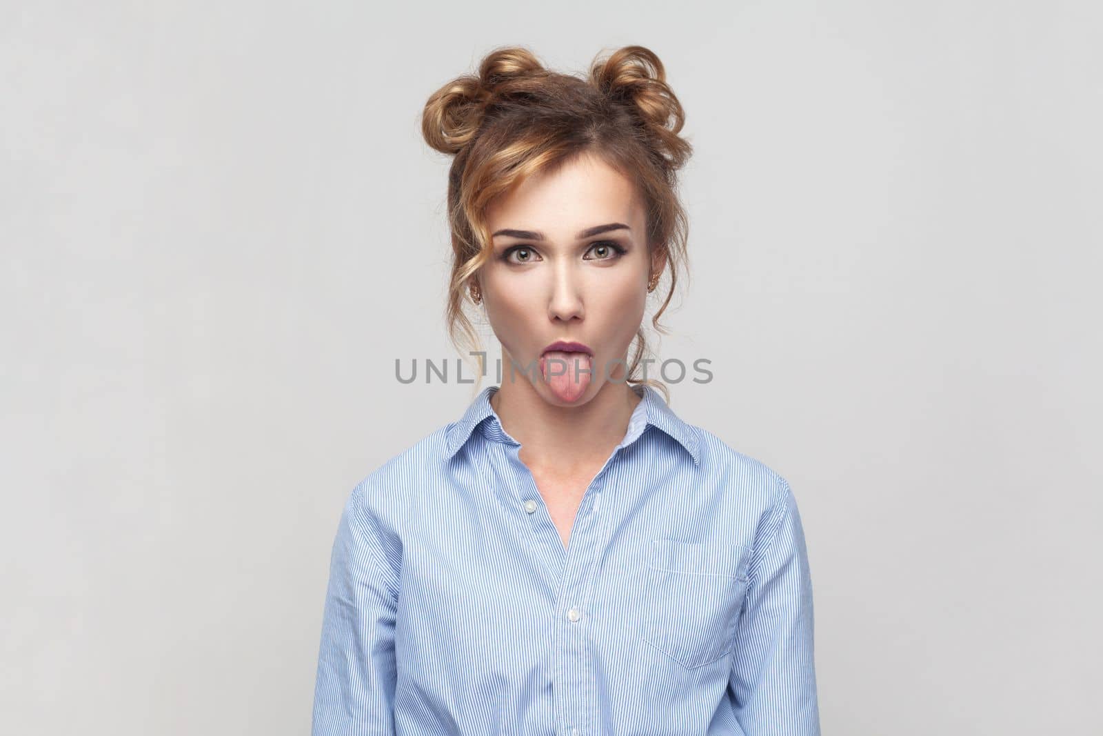 Portrait of naughty comic blonde woman sticking out tongue, misbehaves, demonstrating her disobedience and protest, has funny look, wearing blue shirt. Indoor studio shot isolated on gray background.