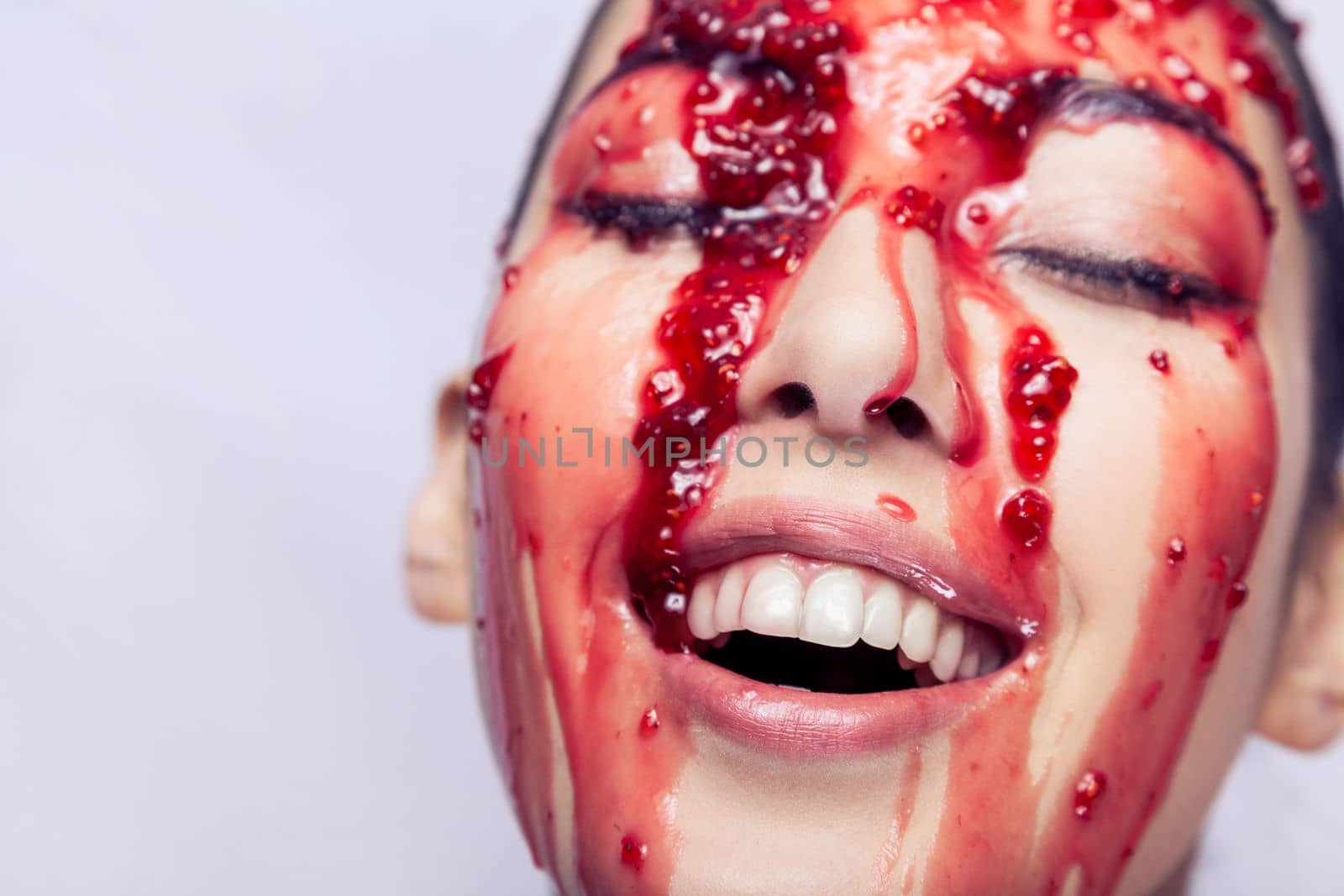 Fashion portrait of woman with closed eyes and smiling, model in raspberry or strawberry jam flowing in the face and the girl enjoying, laughing. Indoor studio shot isolated on gray background.