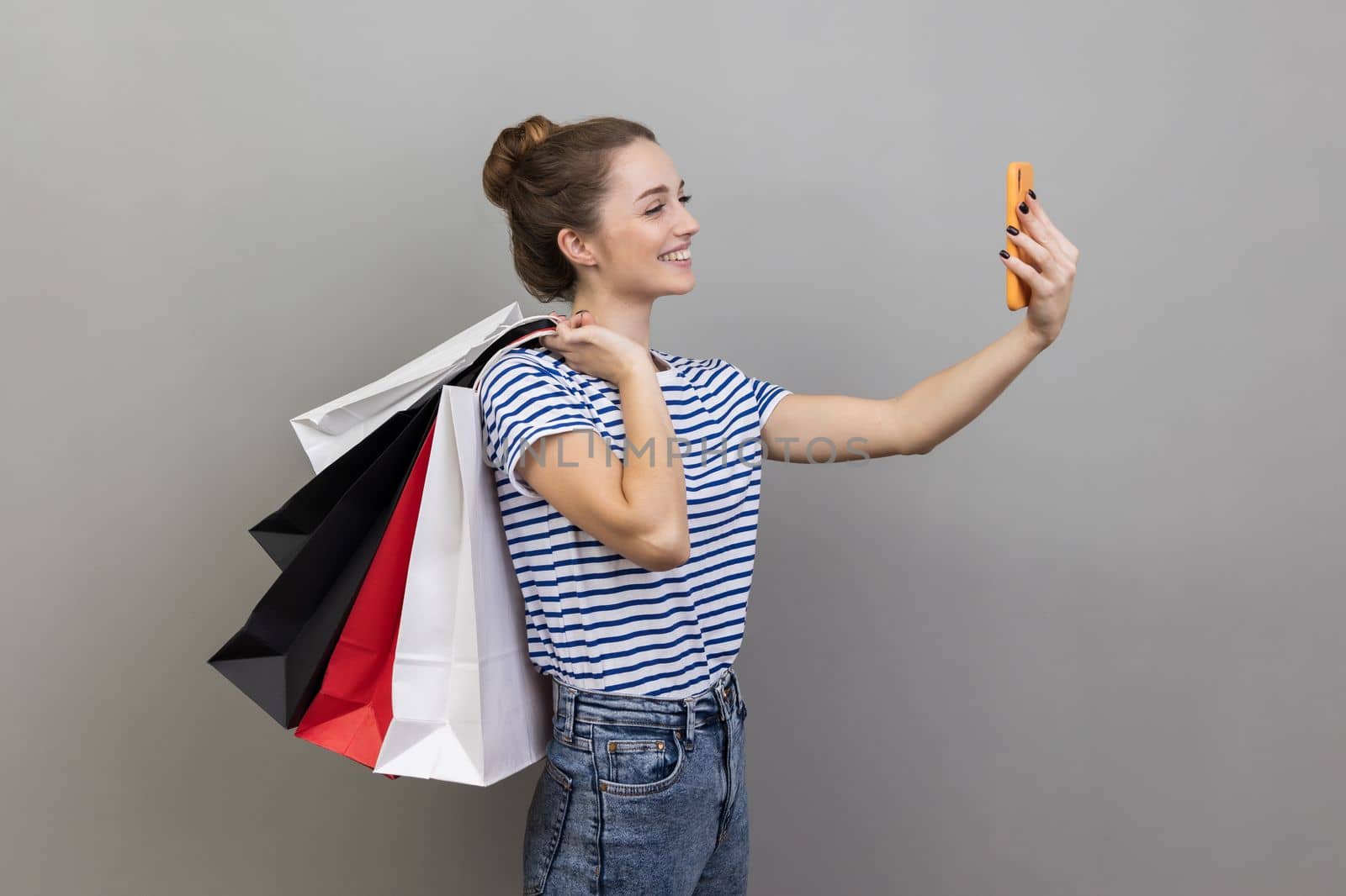 Side view portrait of woman wearing striped T-shirt broadcasting livestream and boasting her purchase, holding shopping, using smartphone. Indoor studio shot isolated on gray background.