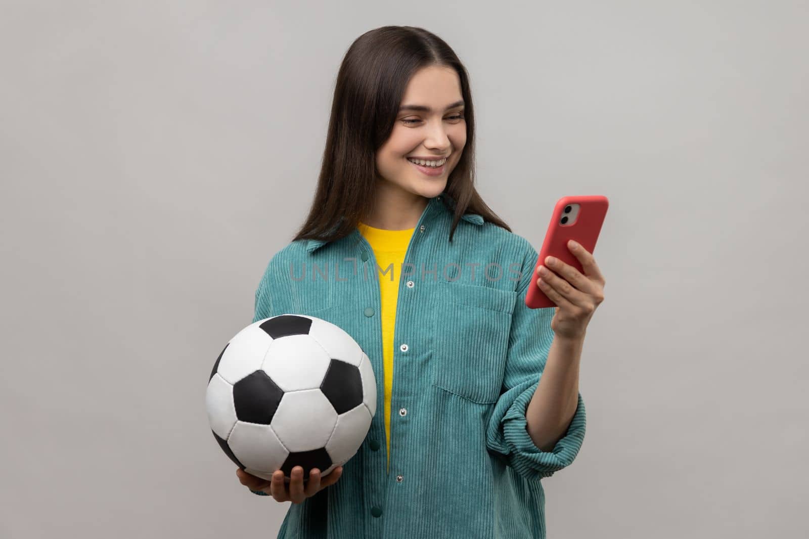 Smiling delighted woman standing holding soccer ball and using smart phone, expressing positive emotions, wearing casual style jacket. Indoor studio shot isolated on gray background.