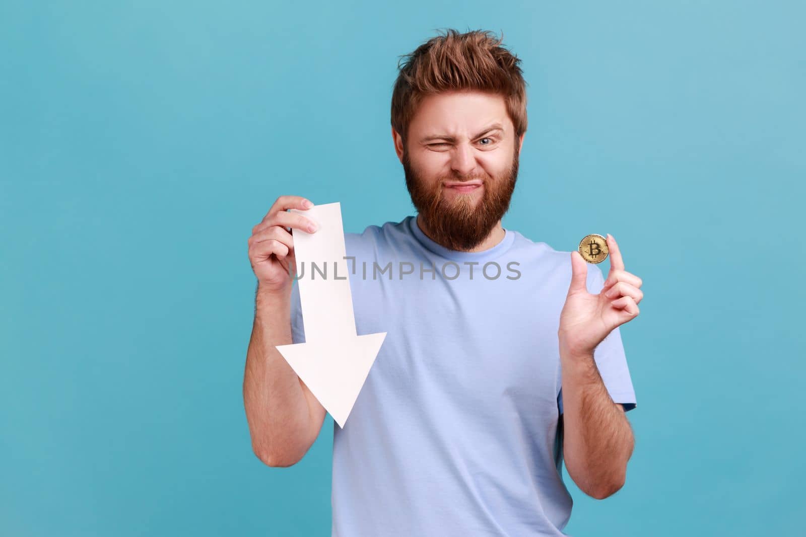 Portrait of upset sad bearded man showing gold bitcoin and arrow pointing down, downgrade of electronic currency, expressing sorrow. Indoor studio shot isolated on blue background.