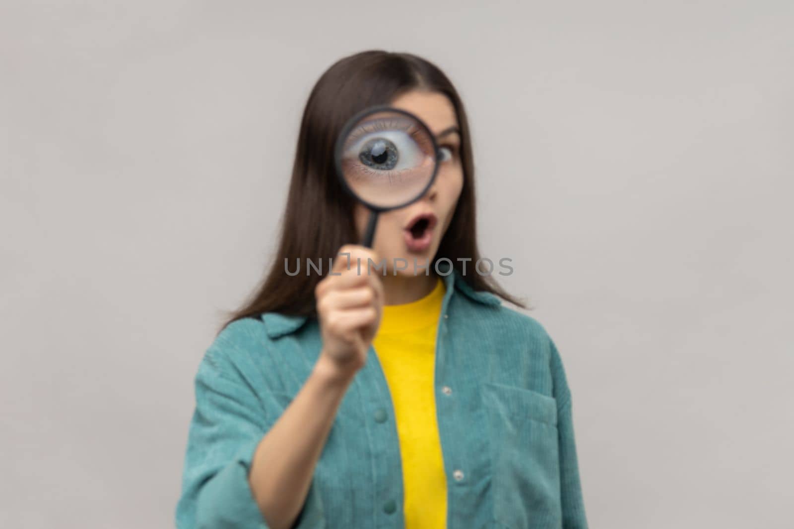Surprised woman standing, holding magnifying glass and looking at camera with big zoom eye, having amazed expression, wearing casual style jacket. Indoor studio shot isolated on gray background.