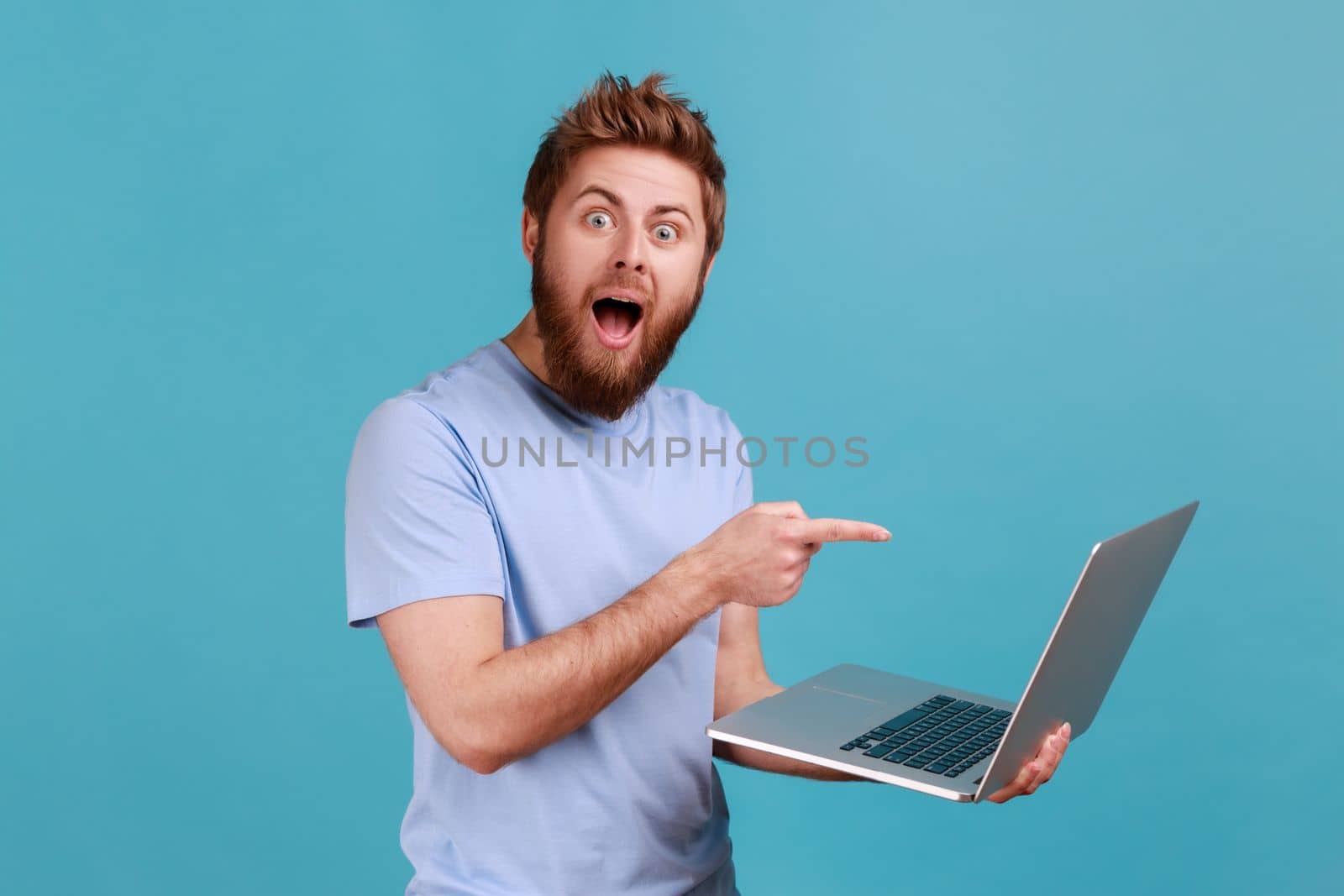 Portrait of shocked bearded man pointing finger at laptop and looking at camera with opened mouth and big eyes, expressing astonishment. Indoor studio shot isolated on blue background.