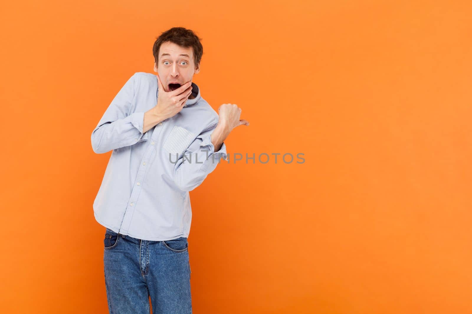 Man looks with jaw dropped out at camera, being horrified to see something, indicates with thumb aside, advertisement area, wearing light blue shirt. Indoor studio shot isolated on orange background.