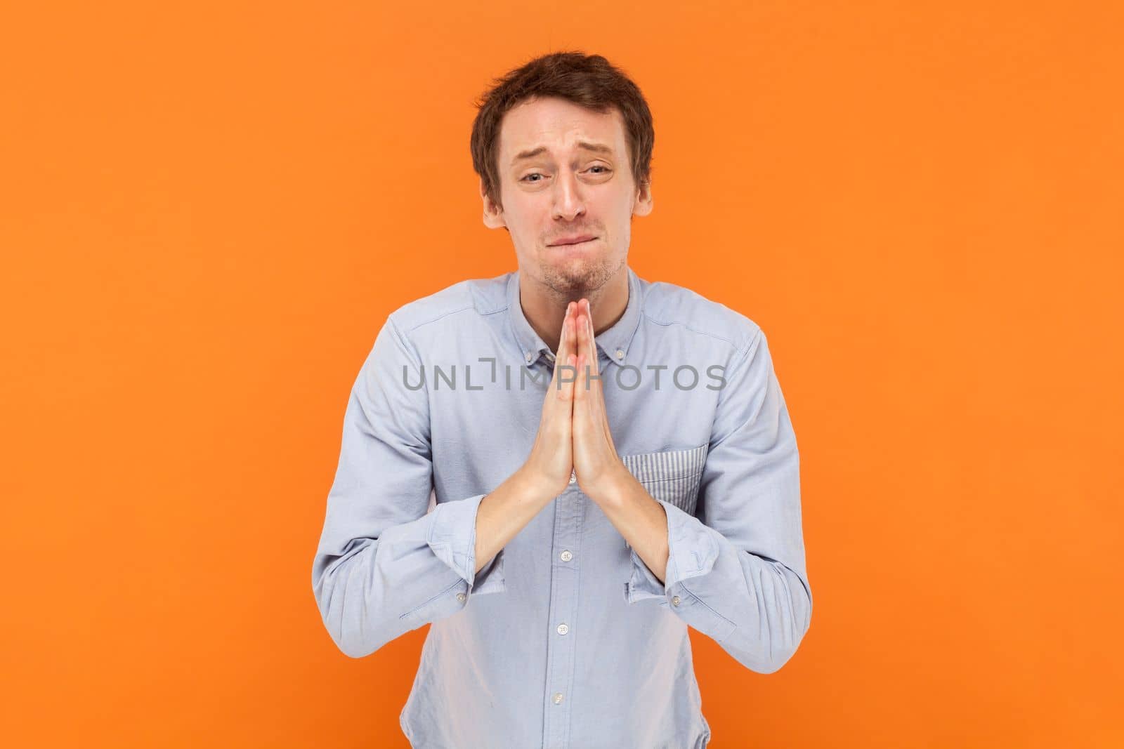 Portrait of hopeful handsome man standing with palms together, asking to forgive, pleading, looking at camera with pout lips, wearing light blue shirt. Indoor studio shot isolated on orange background