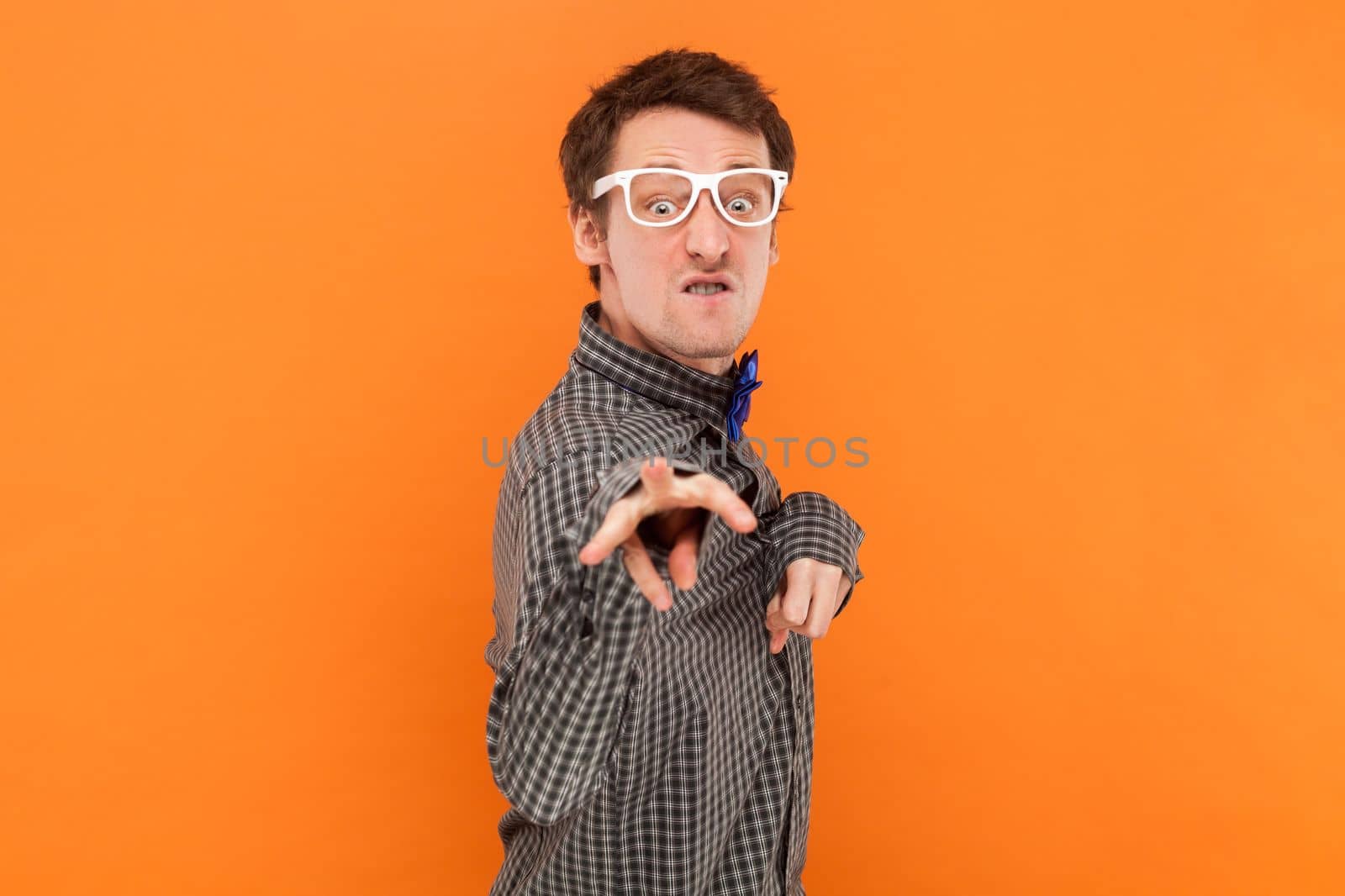Portrait of funny crazy disabled man nerd pointing and looking at camera with clenched teeth, wearing shirt with blue bow tie and white glasses. Indoor studio shot isolated on orange background.