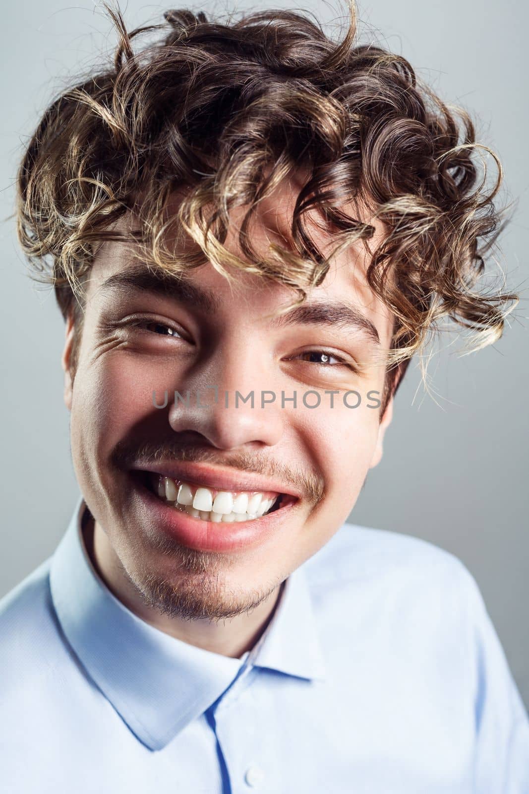 Portrait of happy smiling handsome man with mustache with curly hairstyle, wearing blue shirt, looking at camera, expressing happiness. Indoor studio shot isolated on gray background.
