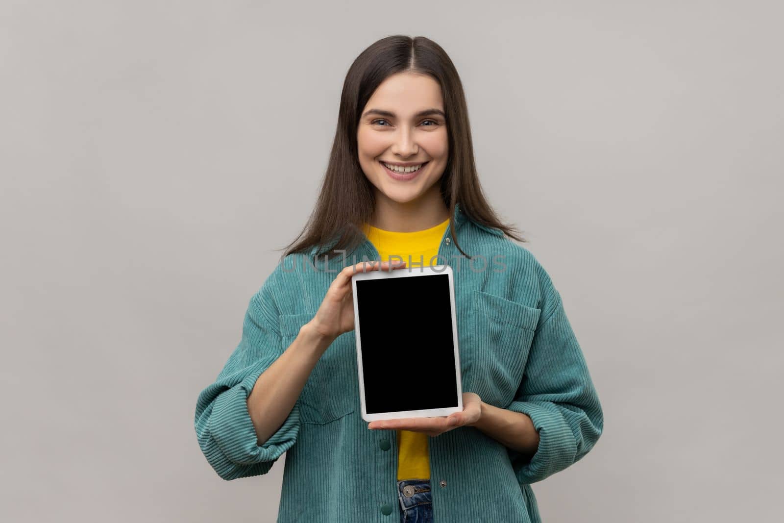 Portrait of positive delighted woman holding showing tablet, looking at camera with smile and happiness, wearing casual style jacket. Indoor studio shot isolated on gray background.