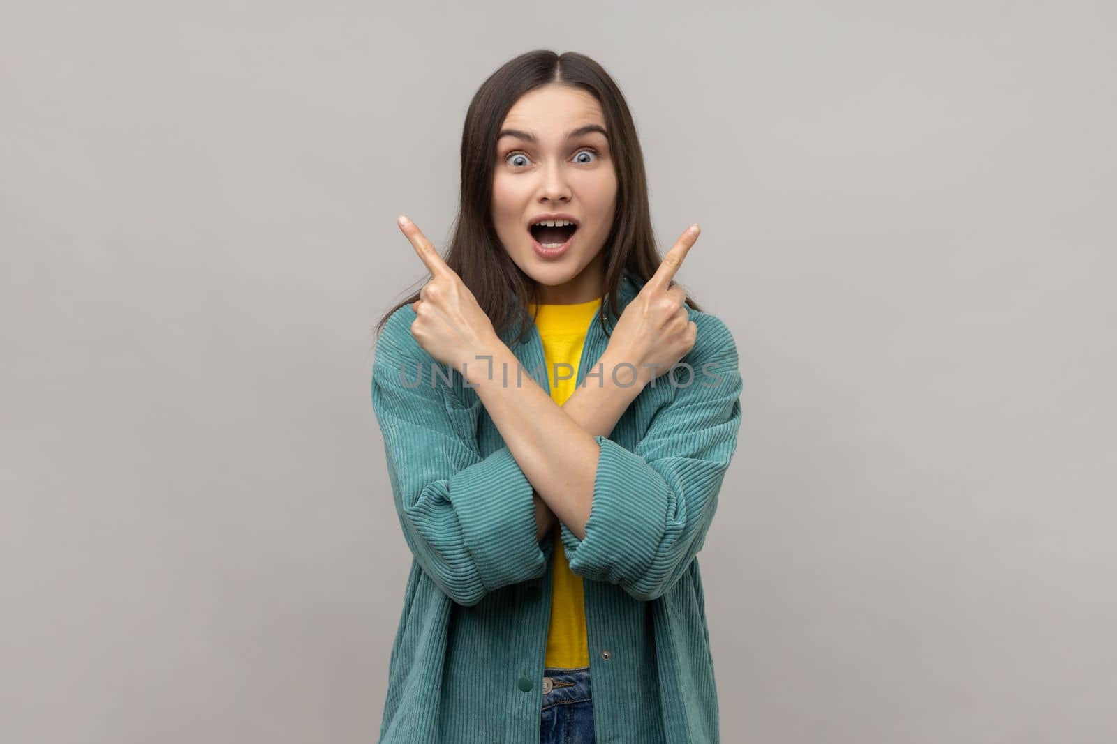 Portrait of surprised woman with dark hair standing with crossed hands and pointing to copy space on both sides, wearing casual style jacket. Indoor studio shot isolated on gray background.