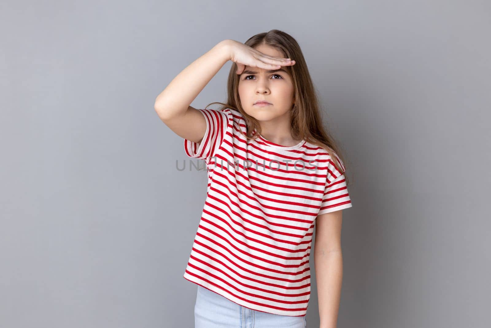 Portrait of little girl wearing striped T-shirt holding palm over her eyes, looking into distance with attentive view, watching horizon, far future. Indoor studio shot isolated on gray background.