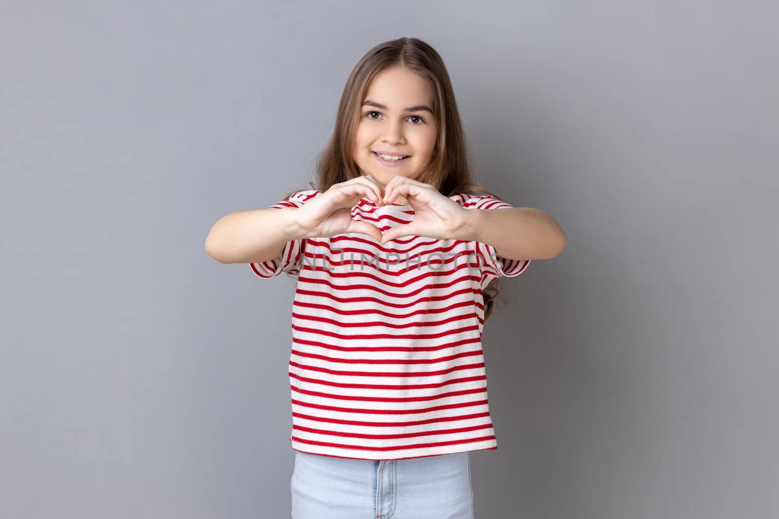 Portrait of little girl wearing striped T-shirt holding hands in shape of heart showing romantic gesture, love confession, valentines day celebration. Indoor studio shot isolated on gray background.