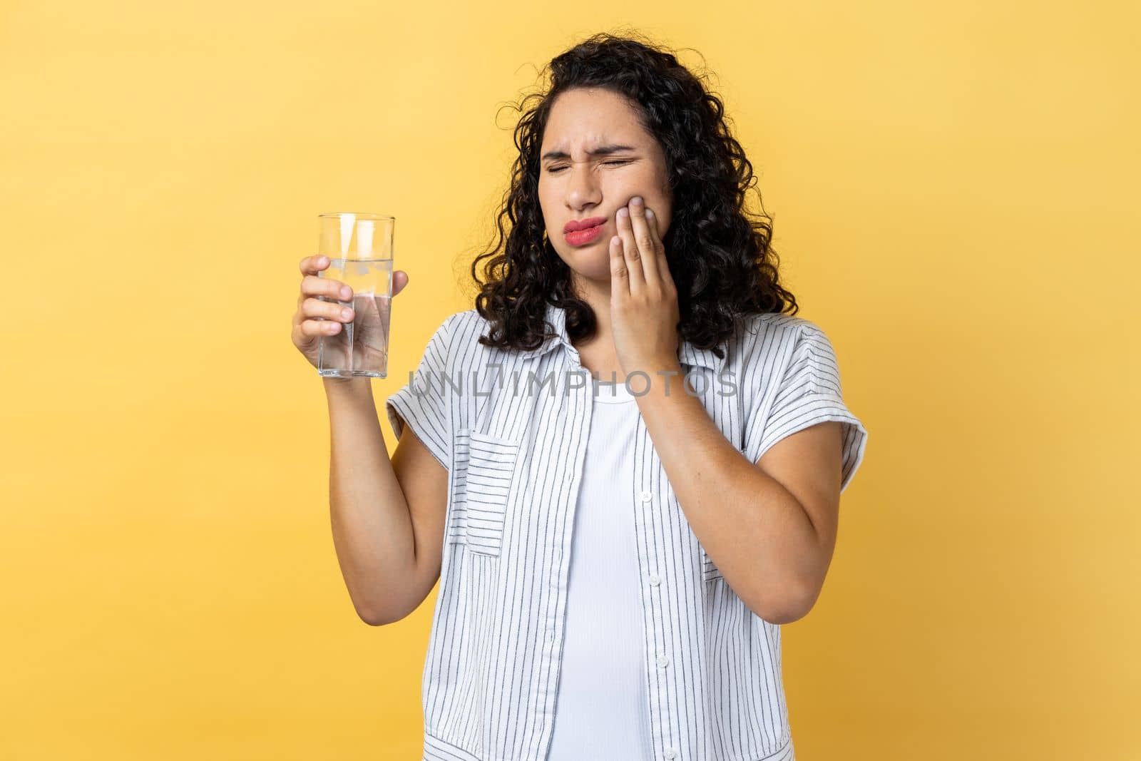 Portrait of attractive woman with dark wavy hair standing and touching his cheek because feeling pain on tooth, after drinking hot tea. Indoor studio shot isolated on yellow background.