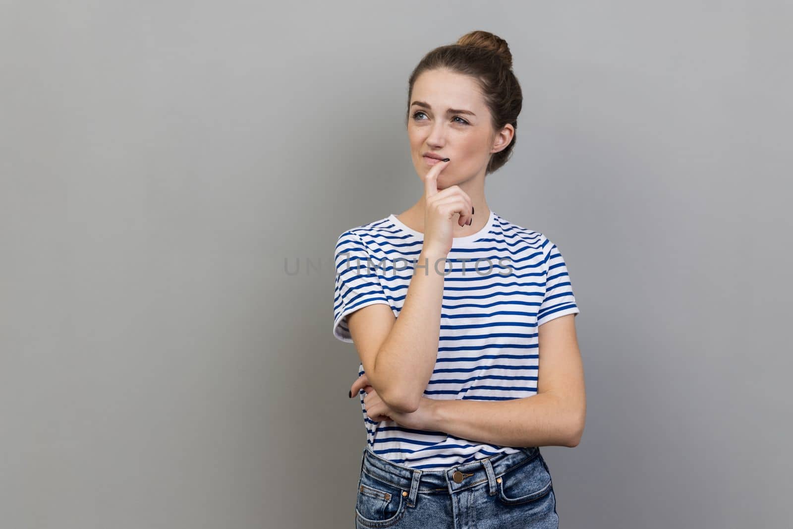 Portrait of woman wearing striped T-shirt pondering serious issues, looking with uncertain hesitant expression, making difficult choice. Indoor studio shot isolated on gray background.